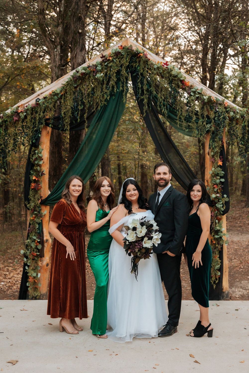A wedding party poses beneath an elegant forest ceremony arch with emerald draping and natural foliage decorations.