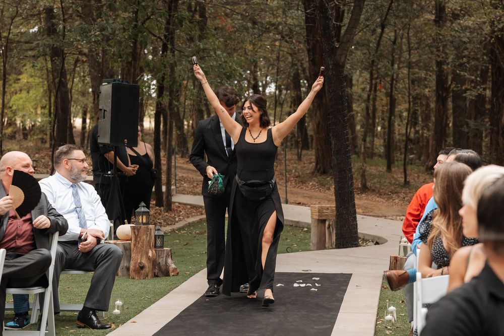 Outdoor wedding ceremony in a wooded area with guests seated in white chairs and a decorated arch at the front.