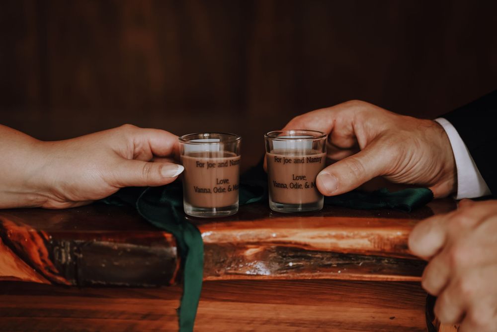 Two hands clink small shot glasses together in a celebratory toast on a wooden surface.