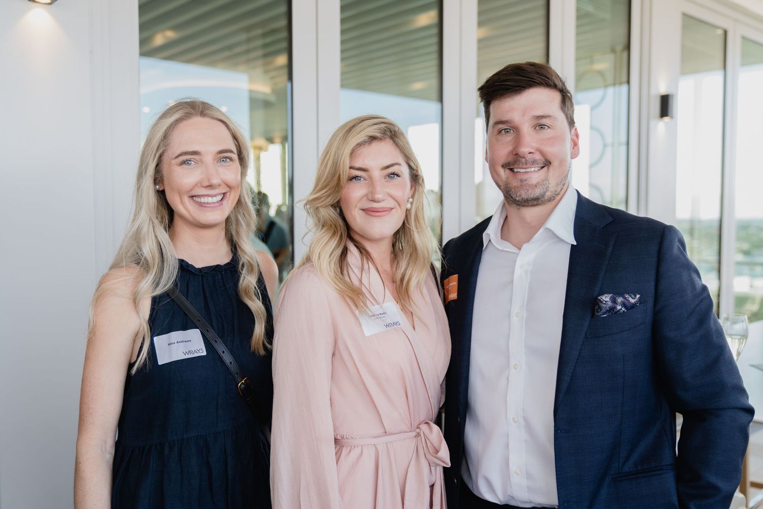 Professional group photo in business attire standing in modern office space with glass windows.