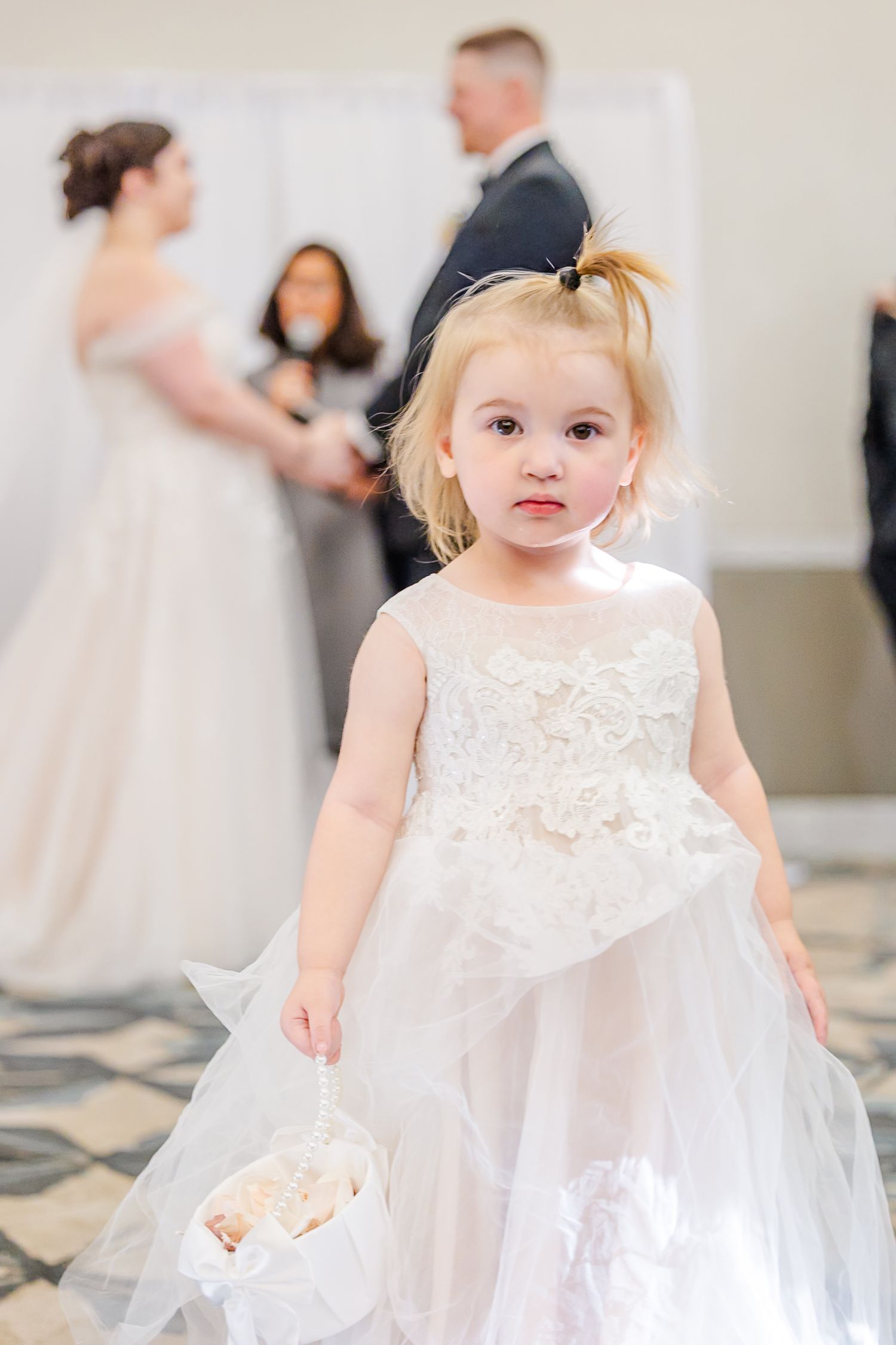 A toddler in a white lace flower girl dress twirls and plays during a formal wedding event.