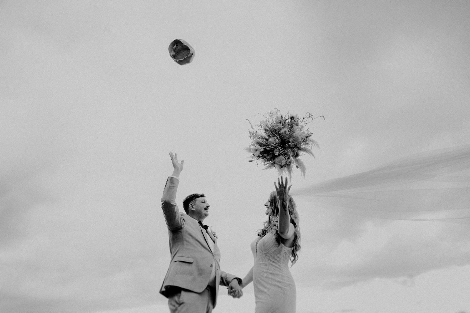 Ein Brautpaar steht Hand in Hand und wirft Strauß und Hut hoch in die Luft. Hochzeit Braunschweig authentische Momente FarmInn Wittingen