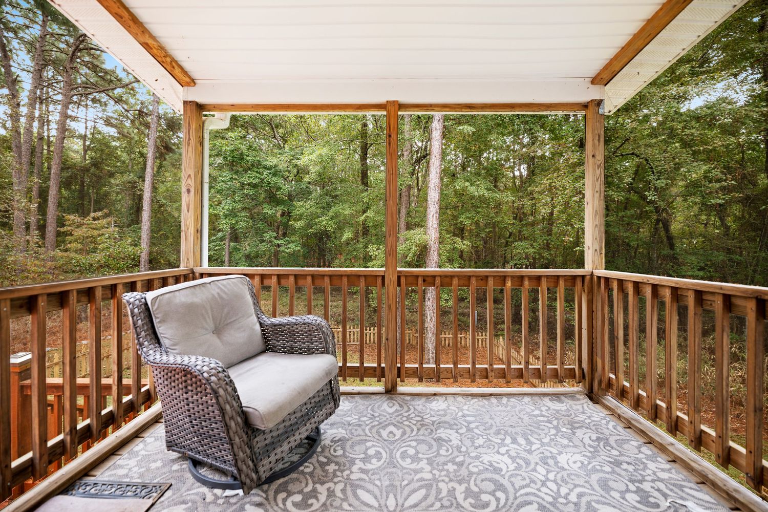 Covered deck with comfortable seating and wooden railings overlooking a wooded area.