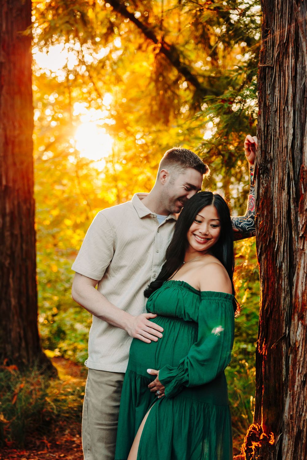 A couple shares an intimate moment among tall redwood trees at sunset with warm golden light filtering through.