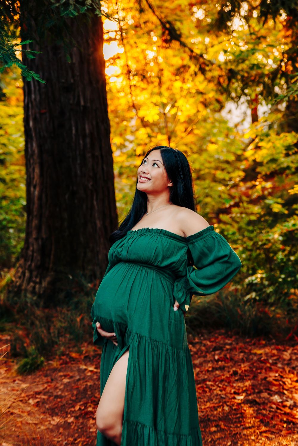 A woman in an emerald green dress poses joyfully against a backdrop of vibrant autumn foliage in a forest setting.