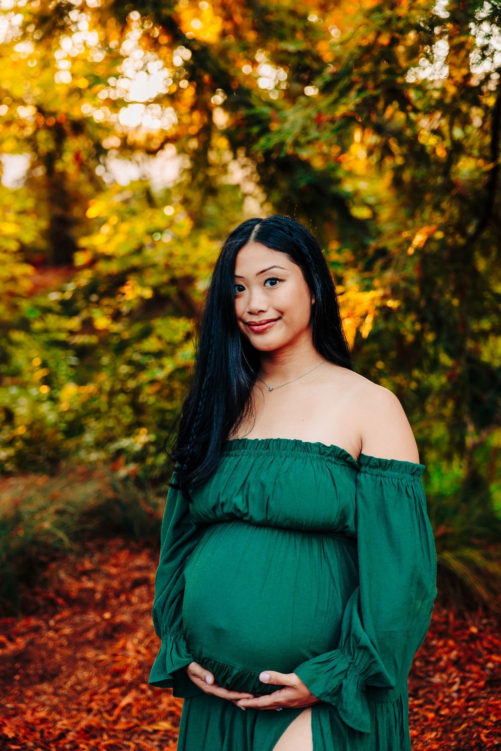A woman in an emerald green off-shoulder dress poses in a forest setting during golden hour.