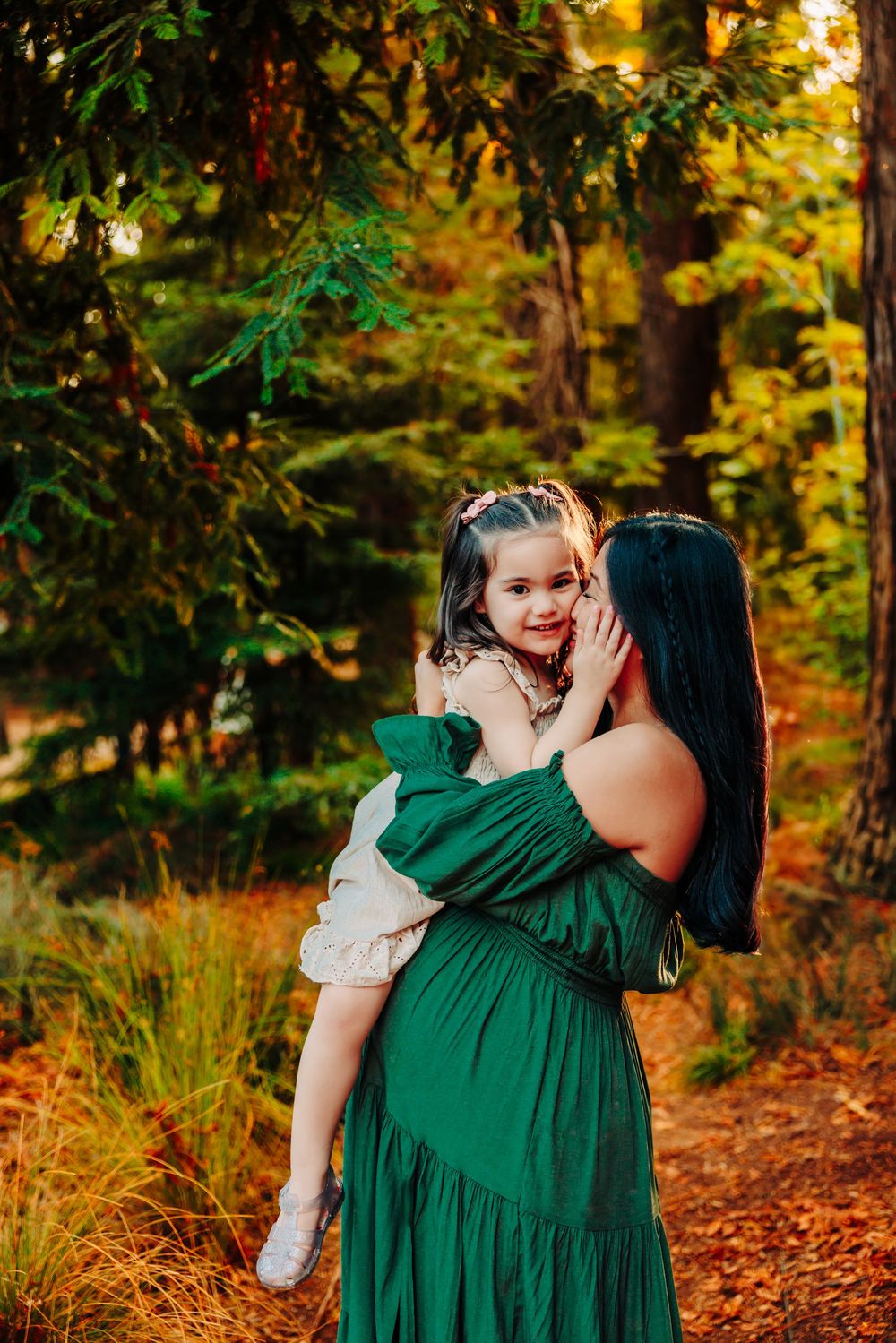 A tender moment between mother and child captured in a sunlit autumn forest with vibrant green and orange colors.