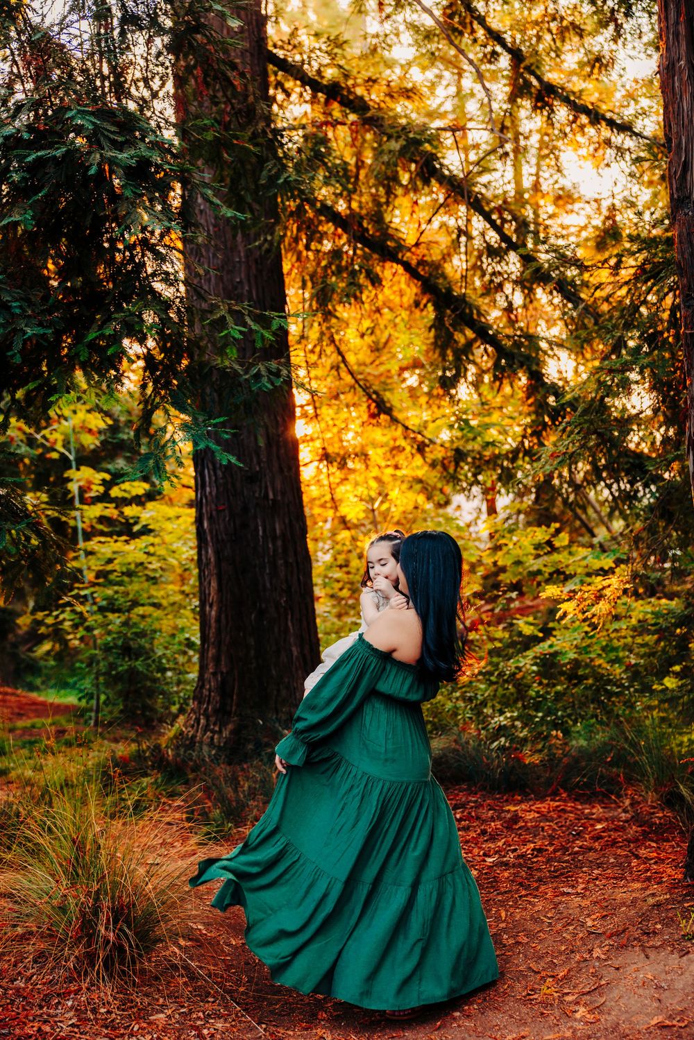 A figure in an emerald green dress poses dramatically against autumn trees at golden hour.