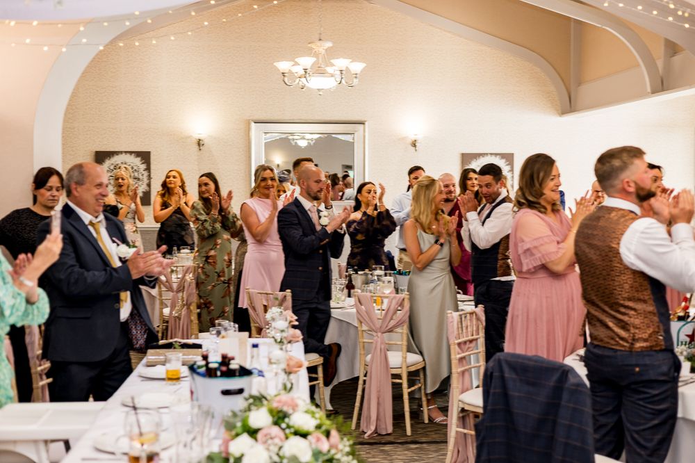 Wedding guests stand and applaud during an elegant reception in a beautifully decorated venue with floral centerpieces.