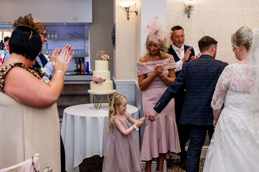 Wedding guests gather around a white tiered cake during the reception celebration.