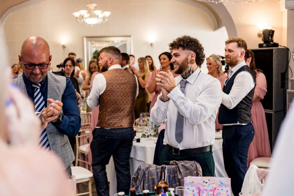 Wedding guests applaud during reception festivities in an elegantly arched venue with chandelier lighting.