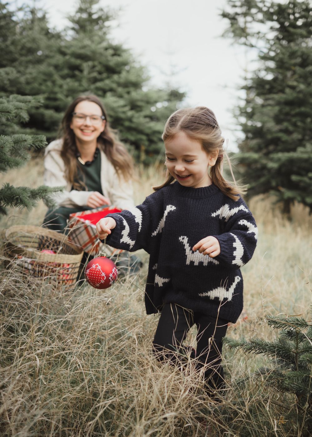 Children decorating a Christmas tree outdoors with ornaments while wearing holiday sweaters in a pine forest setting.