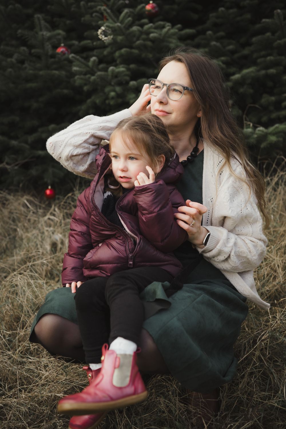 Two people in winter clothing sit together on hay bales surrounded by evergreen trees and holiday decorations.