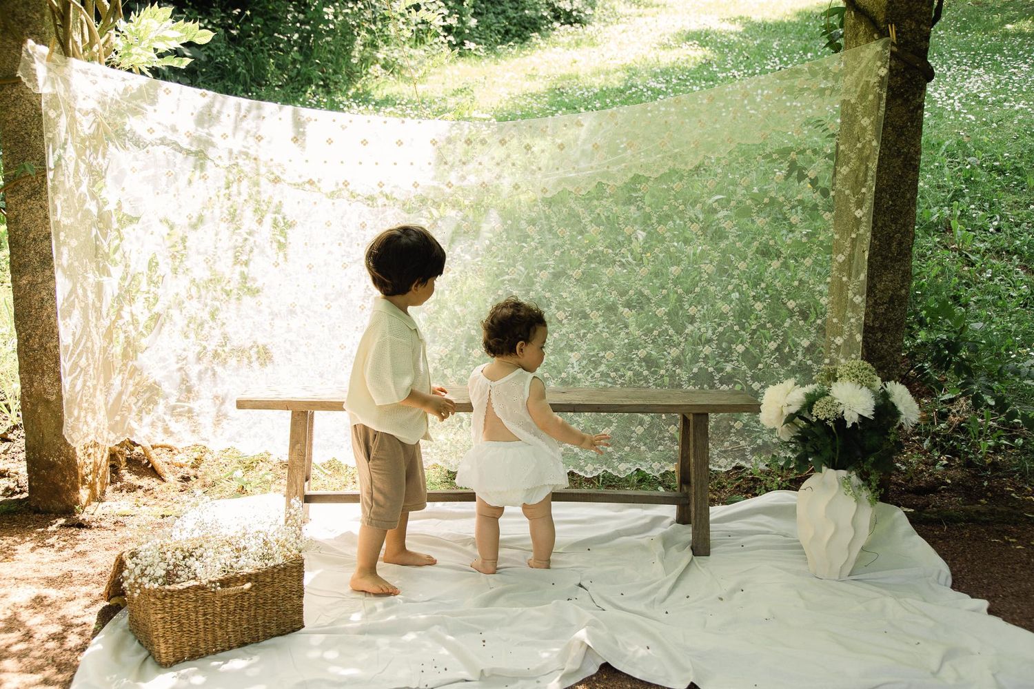 Two young toddler children standing together outdoors under a lace canopy in a garden setting.
