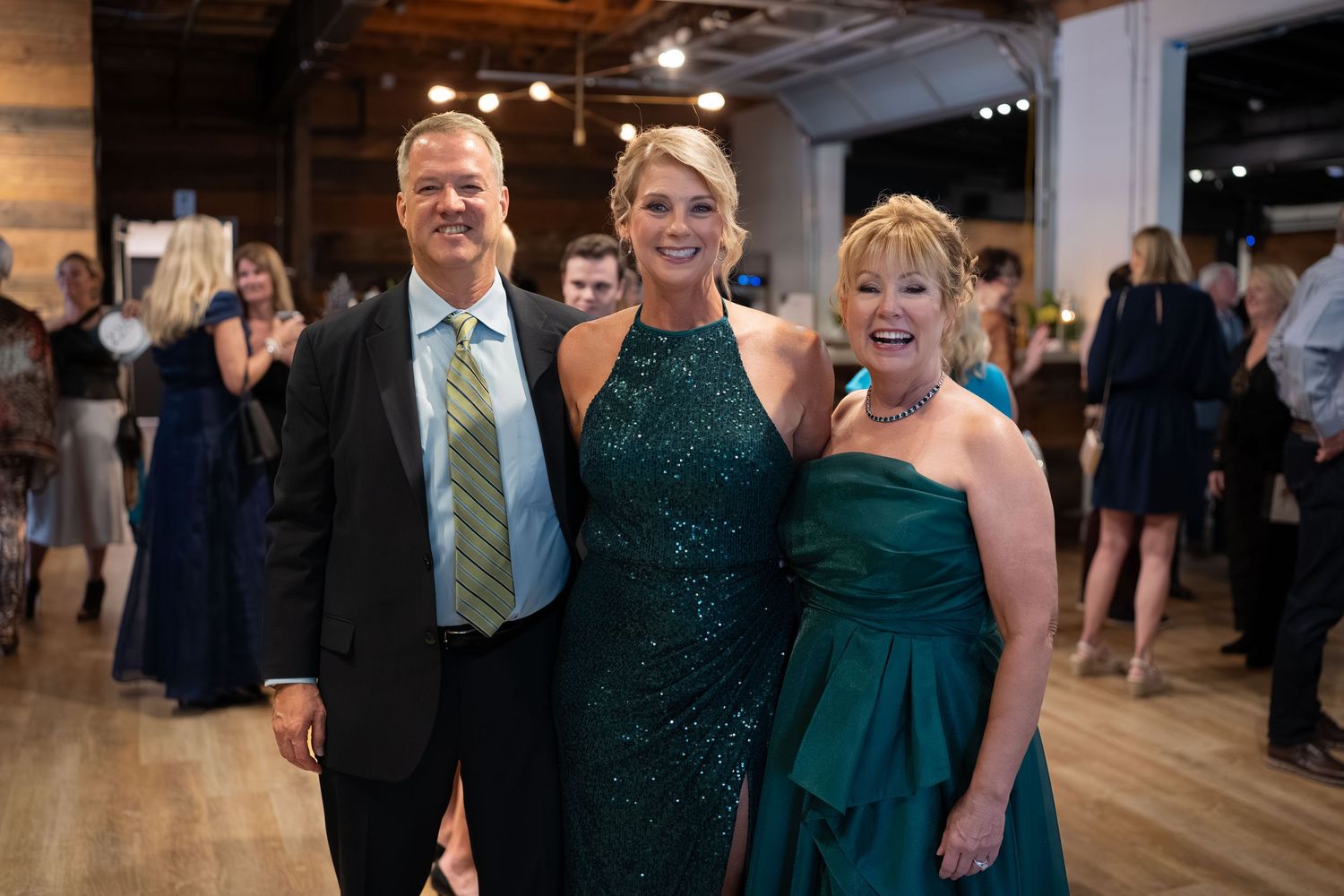 Wedding guests in formal attire including dark suits and emerald green dresses gather at an elegant indoor reception.