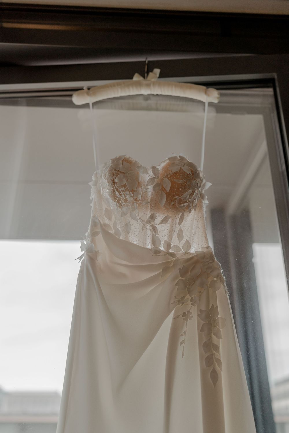 Das Brautkleid der Braut hängt beim Getting Ready am Fenster, damit die Hochzeitsfotografin Detailaufnahmen machen kann. Bildlook ist modern