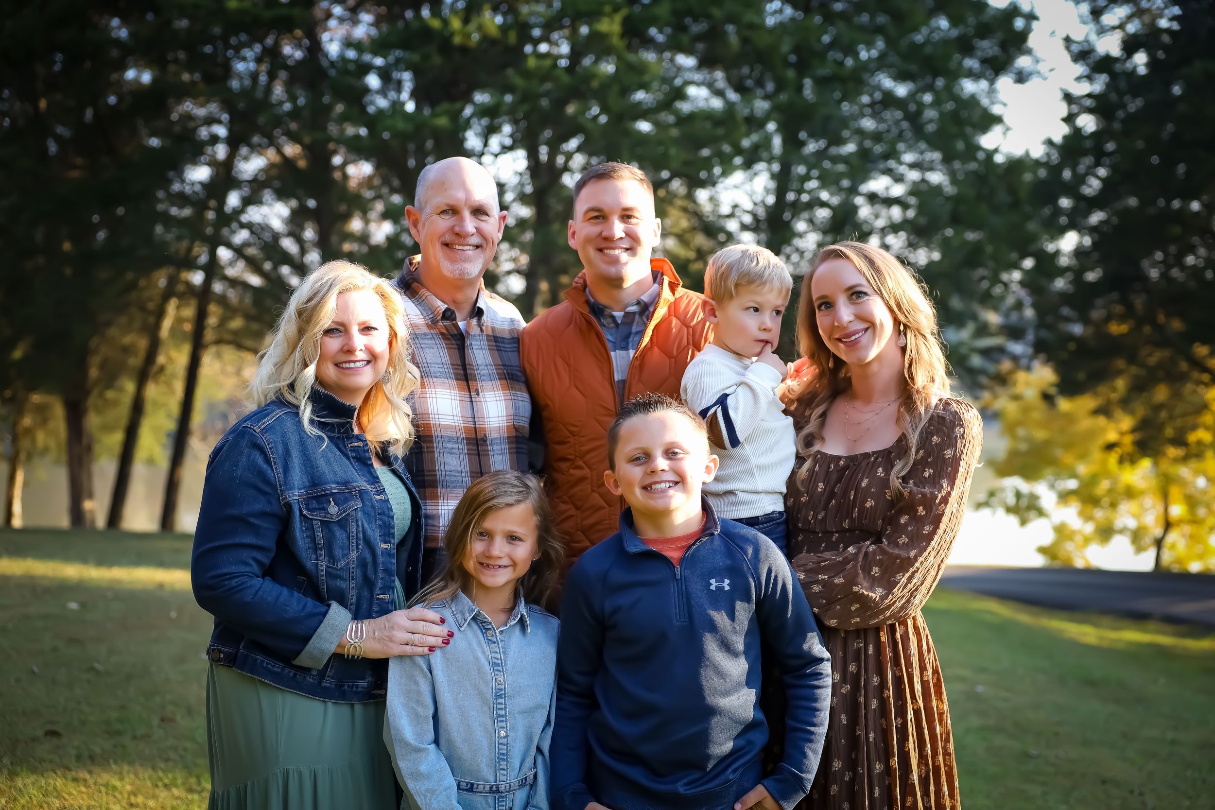 Multi-generational family portrait outdoors in fall clothing with trees in background.