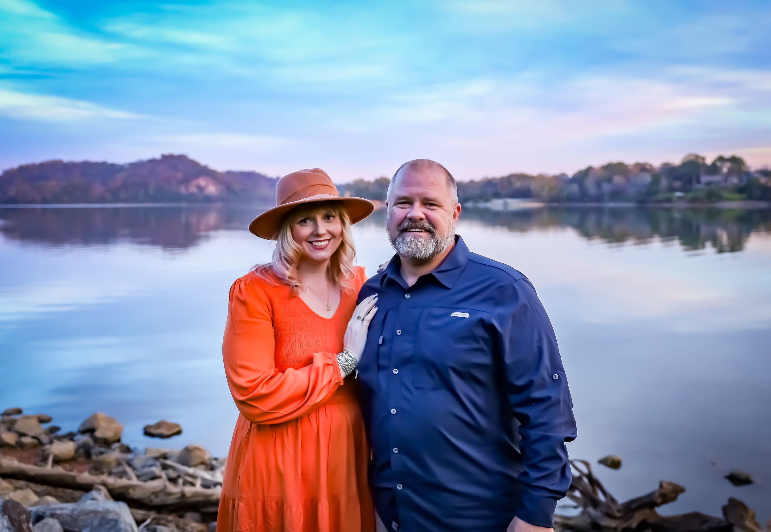 Couple poses by scenic lake at sunset wearing orange dress and navy shirt.