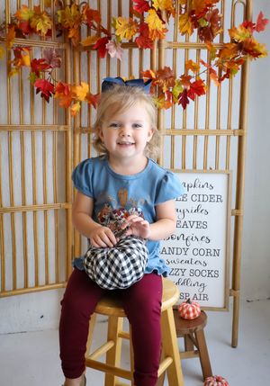 Child in blue shirt sits on wooden stool with autumn decorations and letter board.