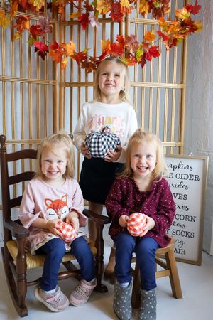 Children sit on wooden chairs decorated with autumn leaves holding small orange pumpkins.