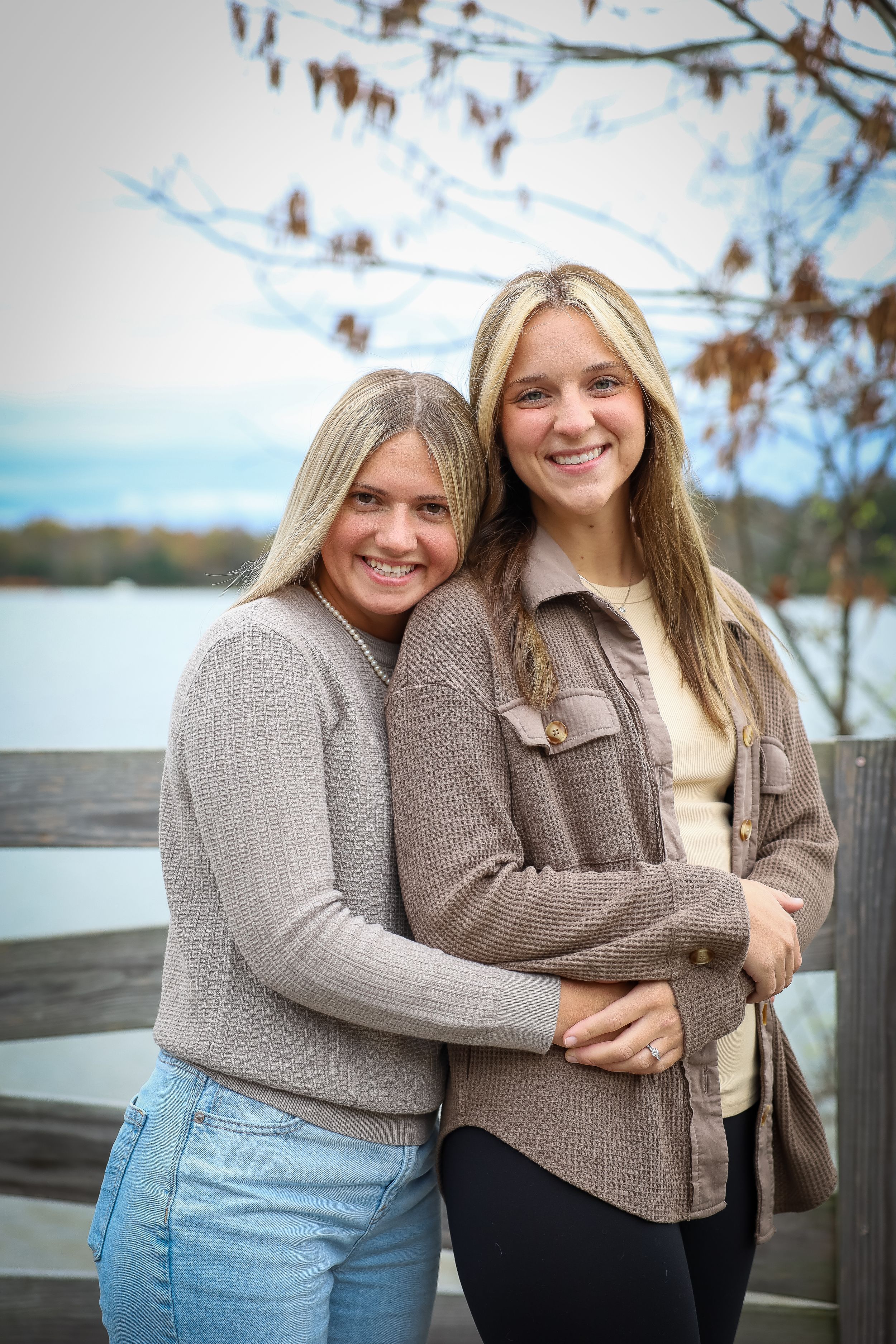 Two women in neutral sweaters embrace on dock overlooking autumn lake.