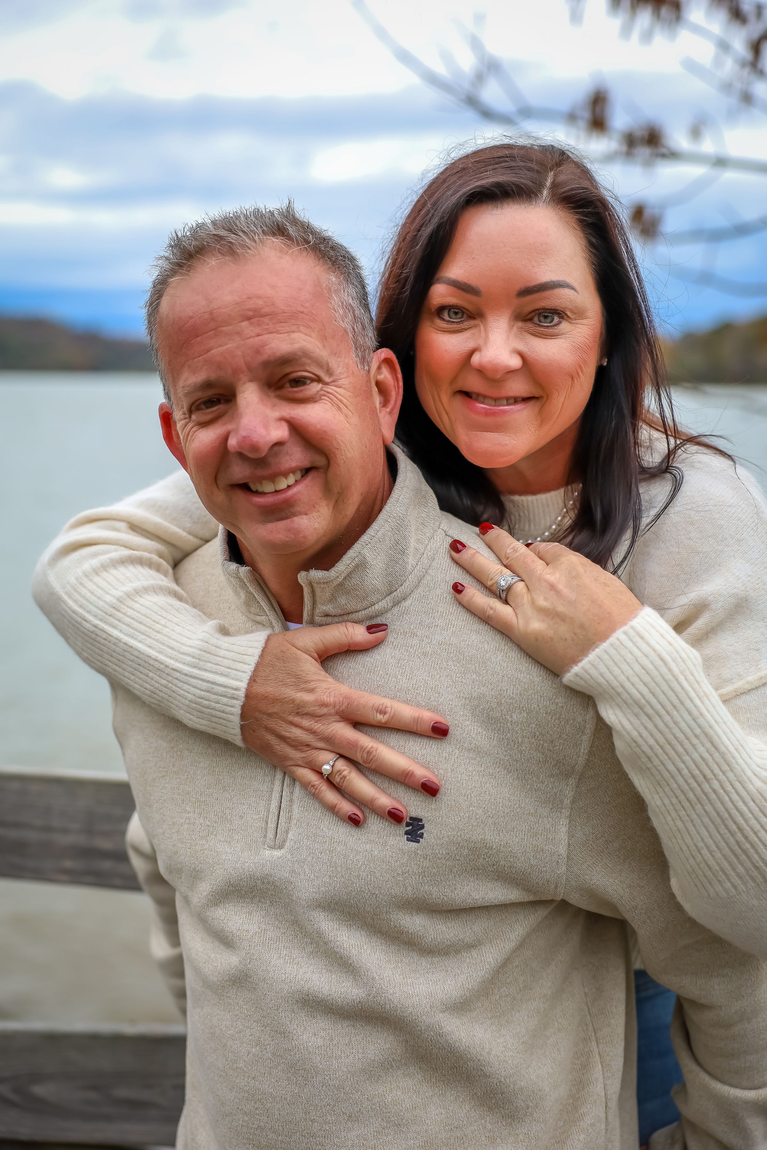 Couple in matching cream sweaters share embrace on lakeside deck.