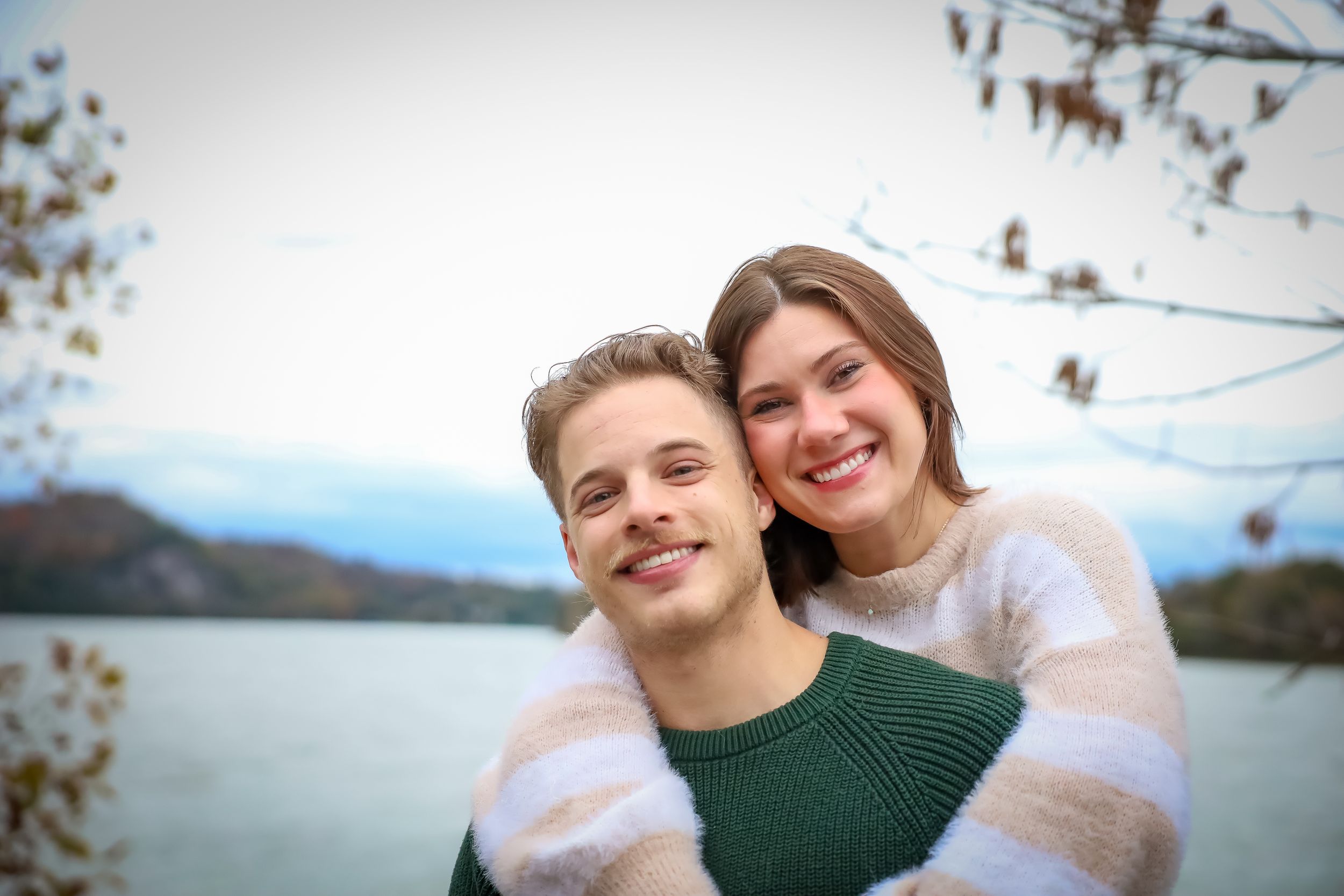Couple in cozy winter wear embracing by lakeside with mountains in background.