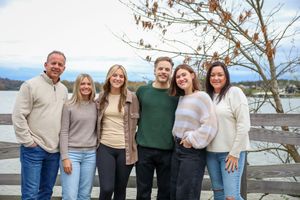 Family group in neutral sweaters poses together on lakeside deck.