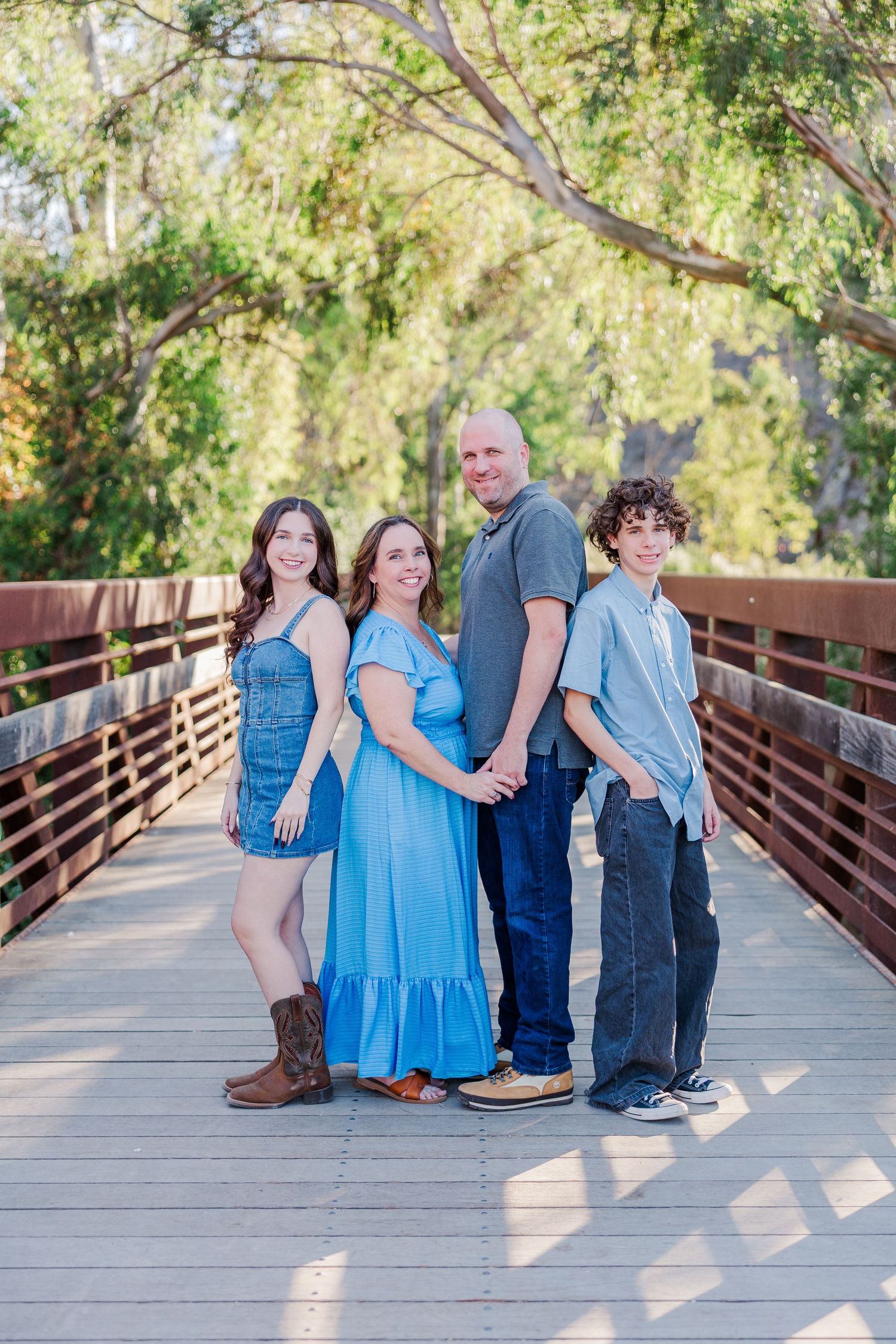 Family wearing coordinating blue and denim outfits sits together on wooden bridge during outdoor photo session.