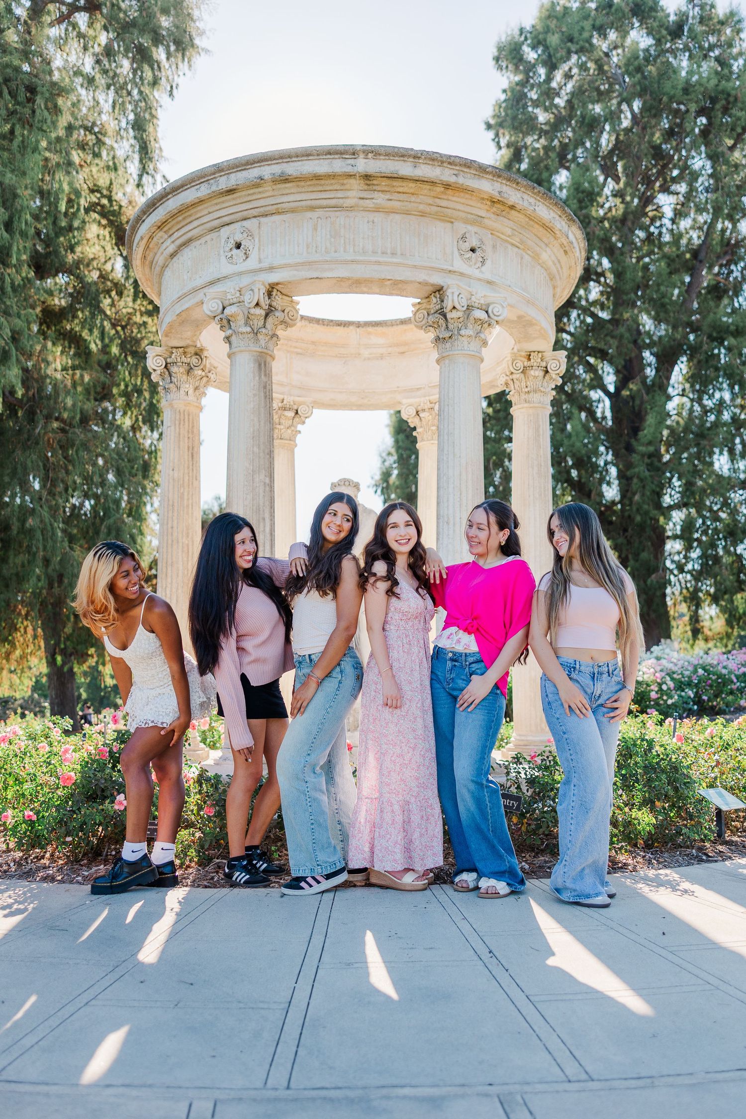 A group of friends in colorful dresses pose together under a white stone gazebo in a garden setting.
