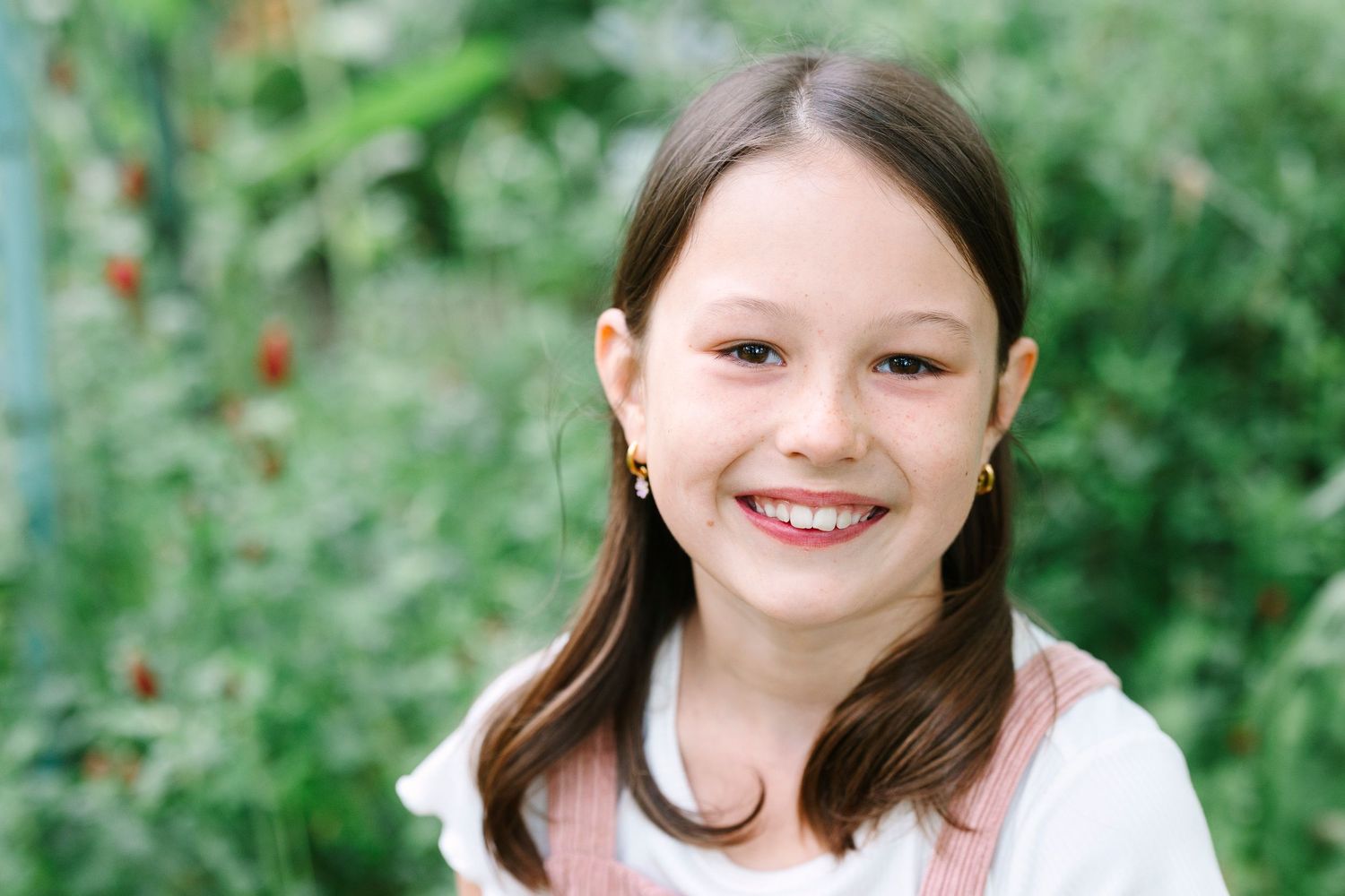 A portrait of a young person in a white top smiling outdoors with lush greenery in the background.