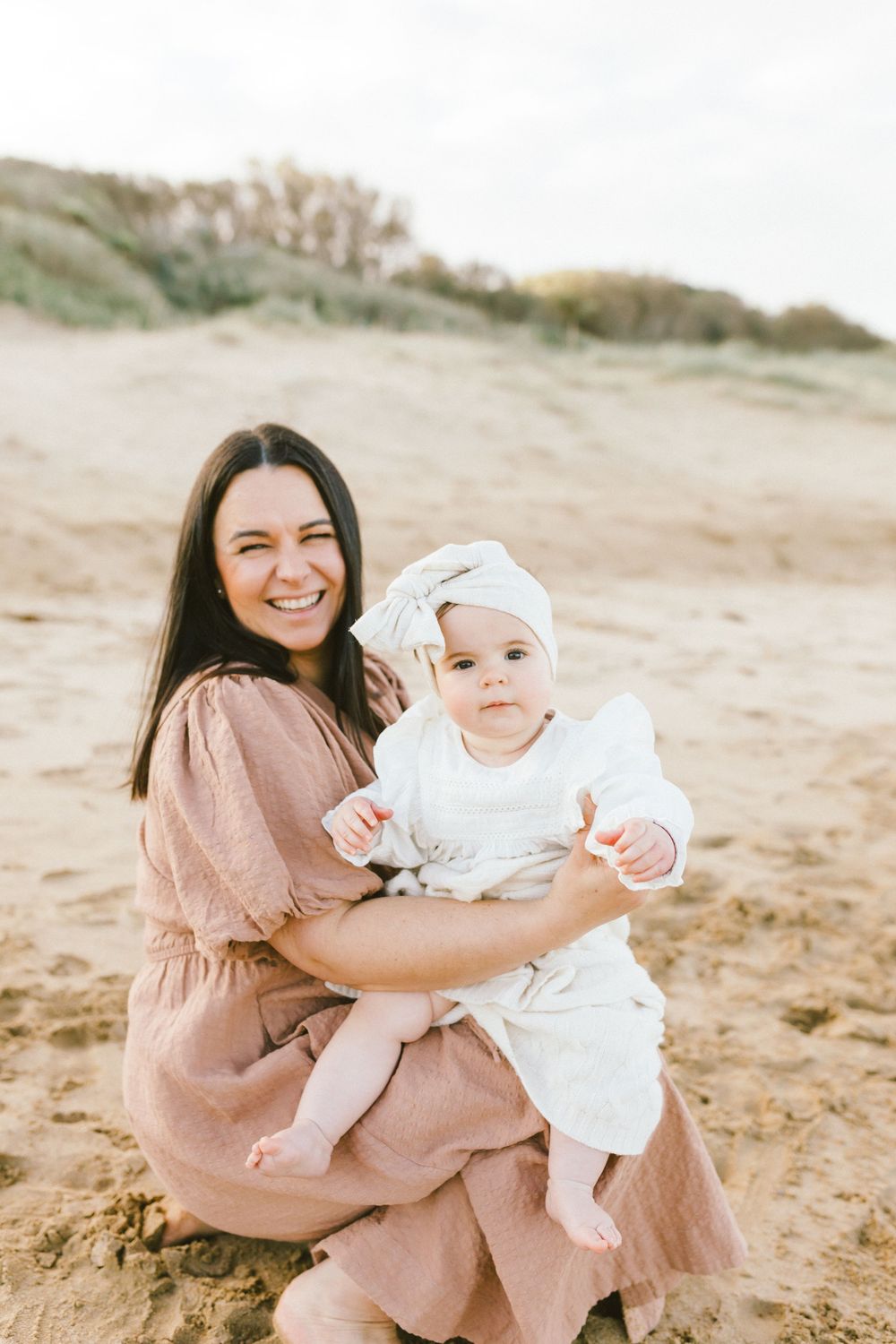 A parent and baby in matching blush tones share tender moments together on sandy terrain during a photo session.