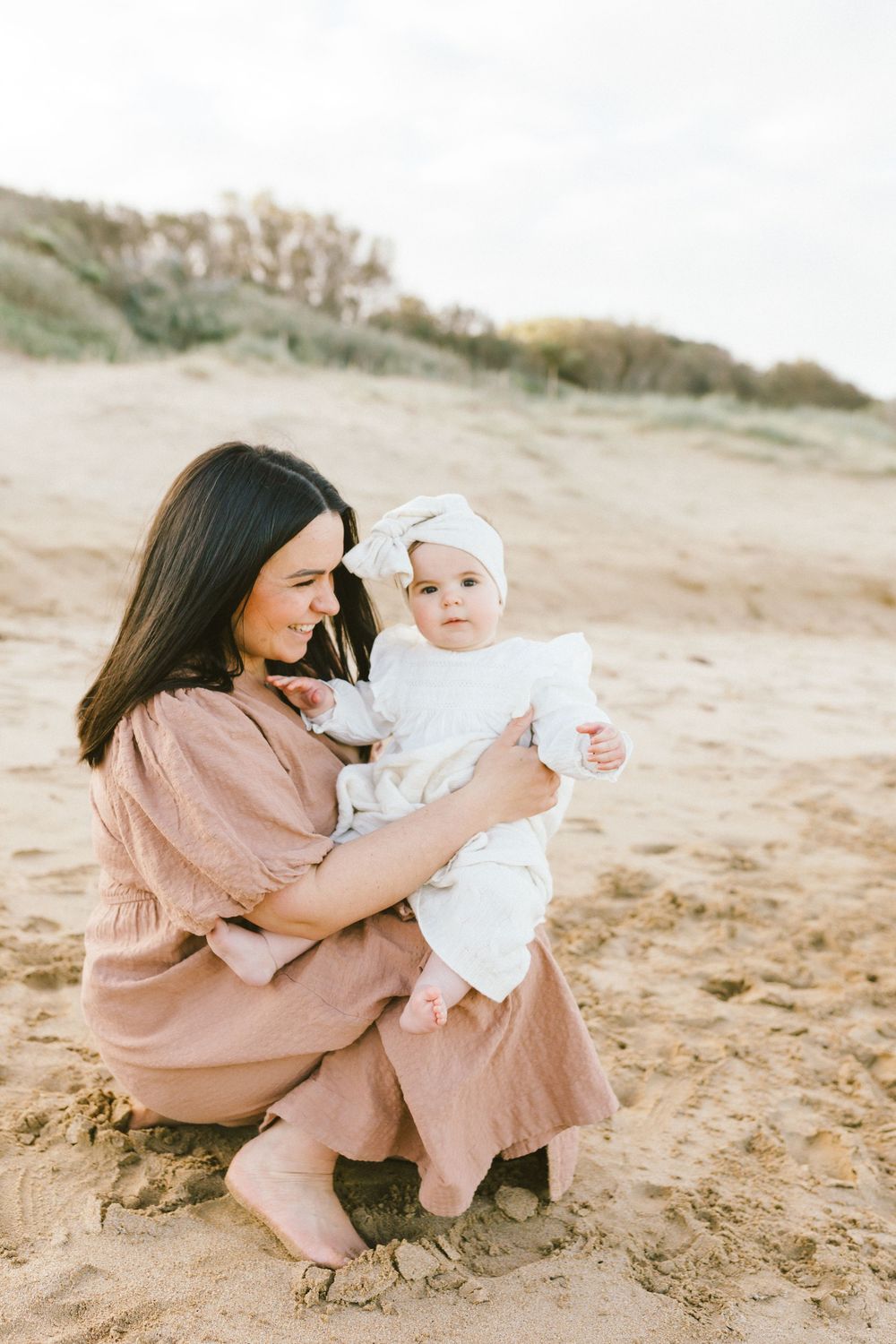 A parent in a pink dress kneels on a sandy beach with a baby wearing white, captured in a series of candid moments.