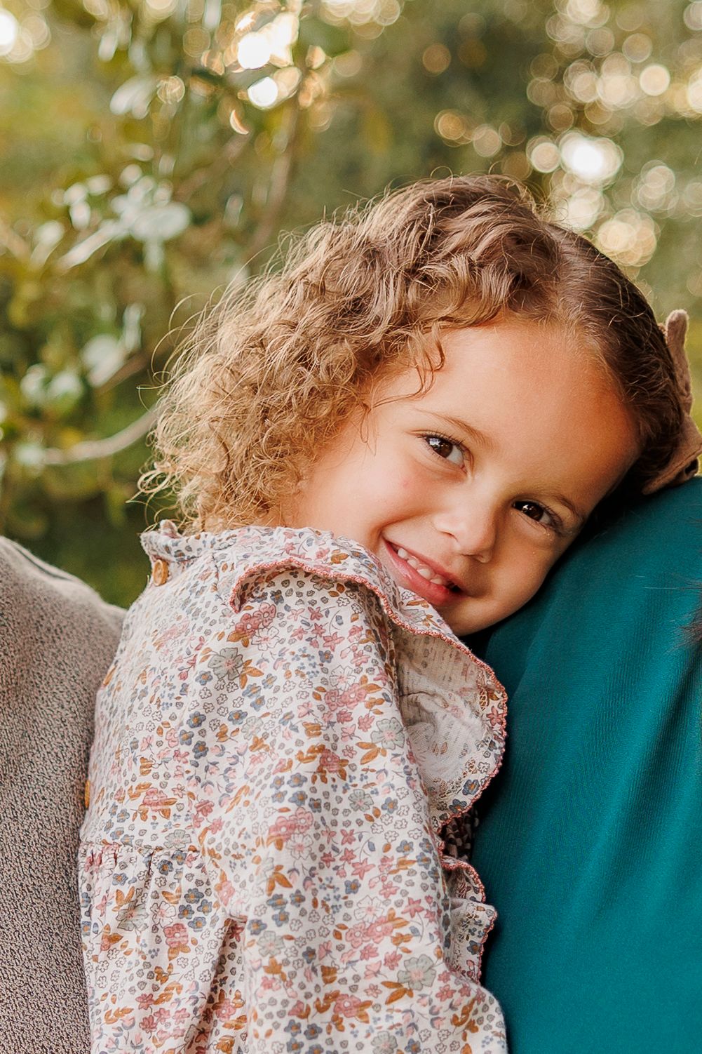 A child in a floral dress leans against a teal-colored surface with natural bokeh lighting in the background.