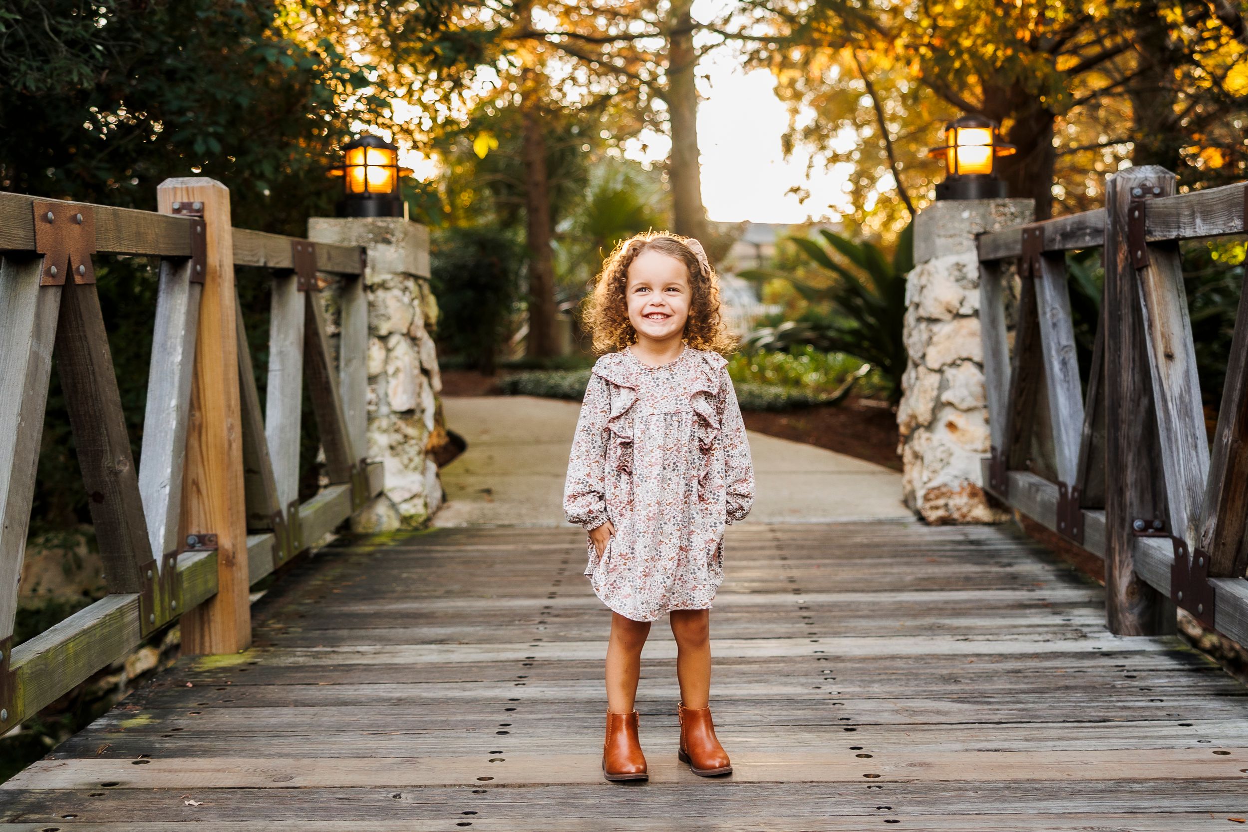 A person in a floral dress stands on a wooden boardwalk surrounded by lush greenery during golden hour.