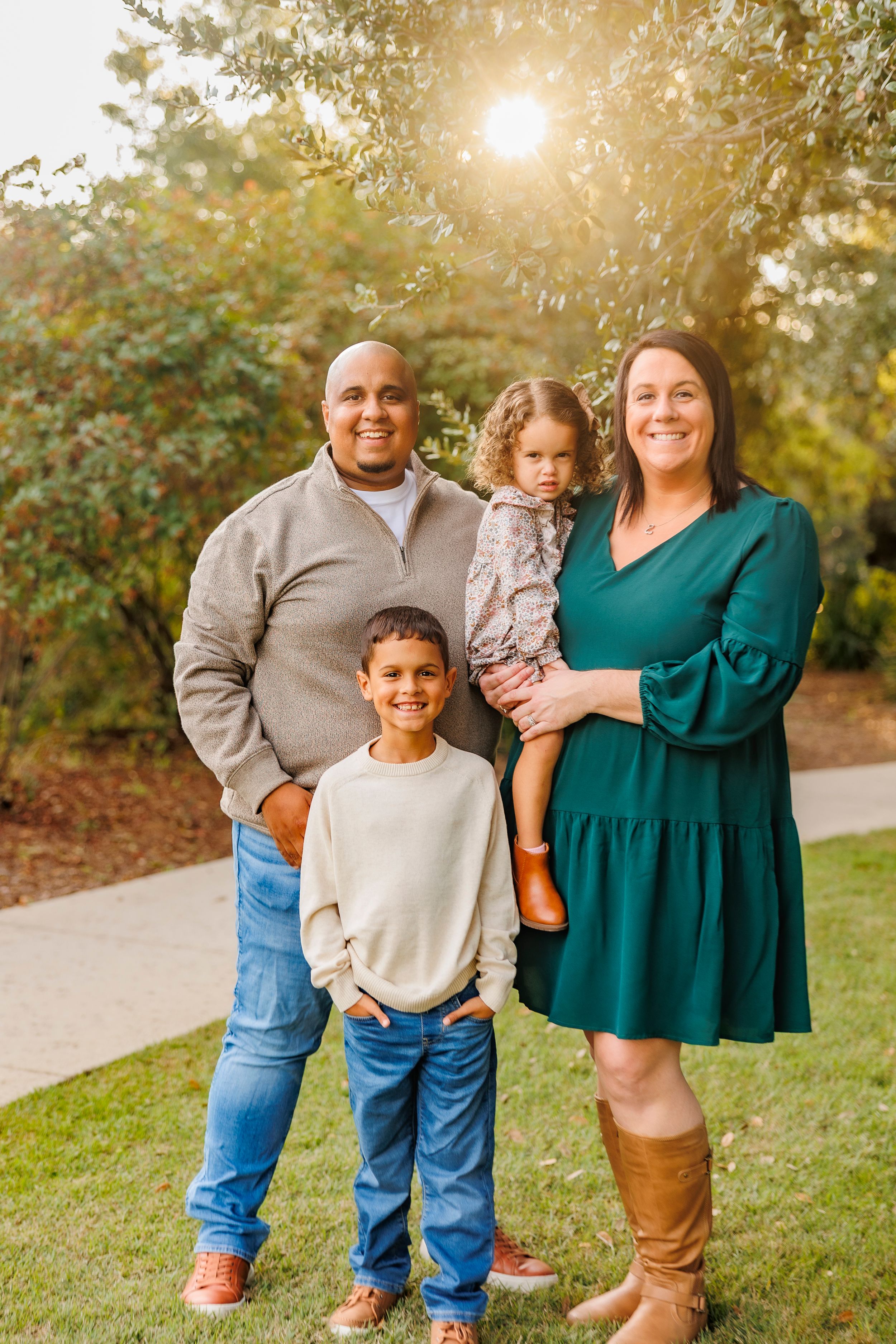 Family posing together outdoors on a path with autumn foliage and warm evening light in the background.