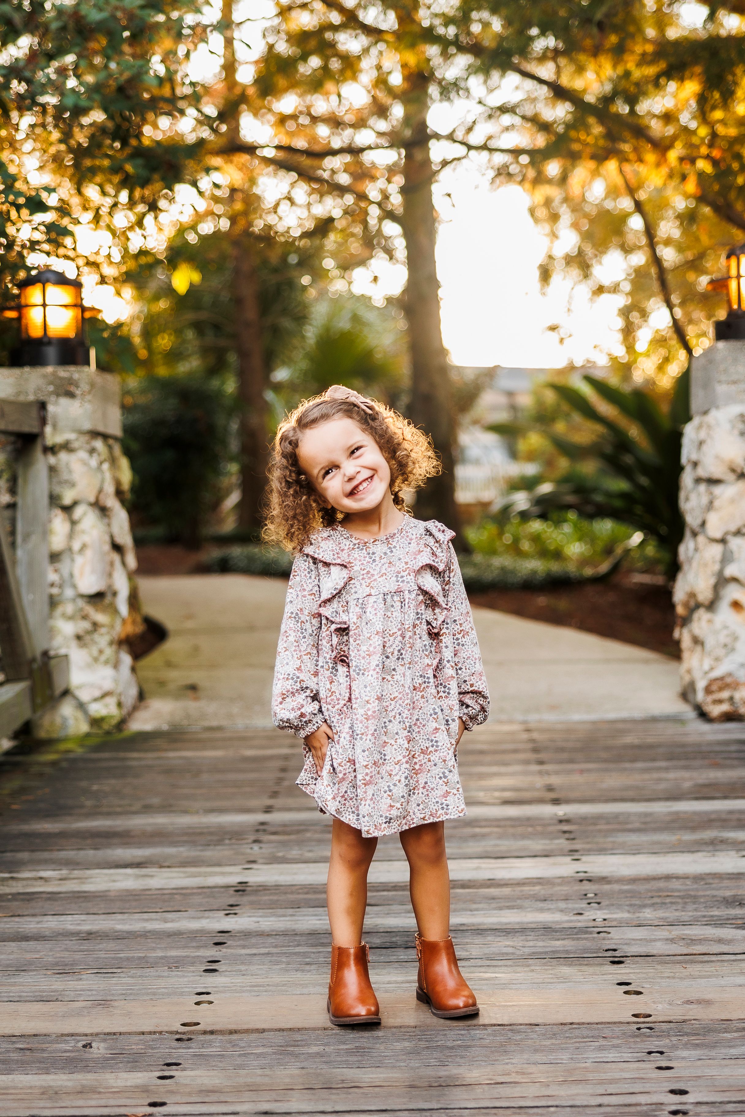 A child in a floral dress and brown boots plays on a wooden boardwalk at sunset among stone pillars and trees.