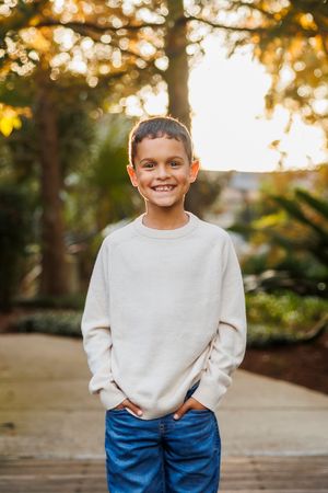 Child in white sweater and blue jeans poses on outdoor walkway during golden hour.