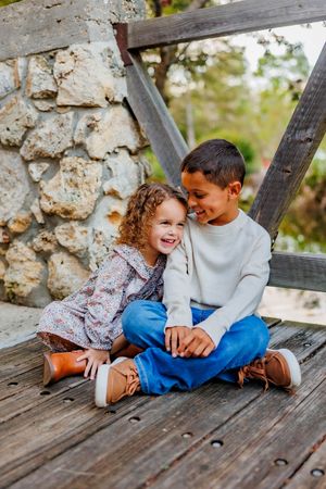 Two children share a joyful moment sitting together on a wooden bridge deck near a stone wall.