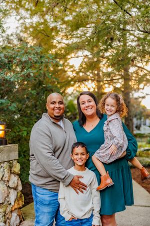 A happy family of four poses together outdoors at sunset with trees and stone wall in the background.