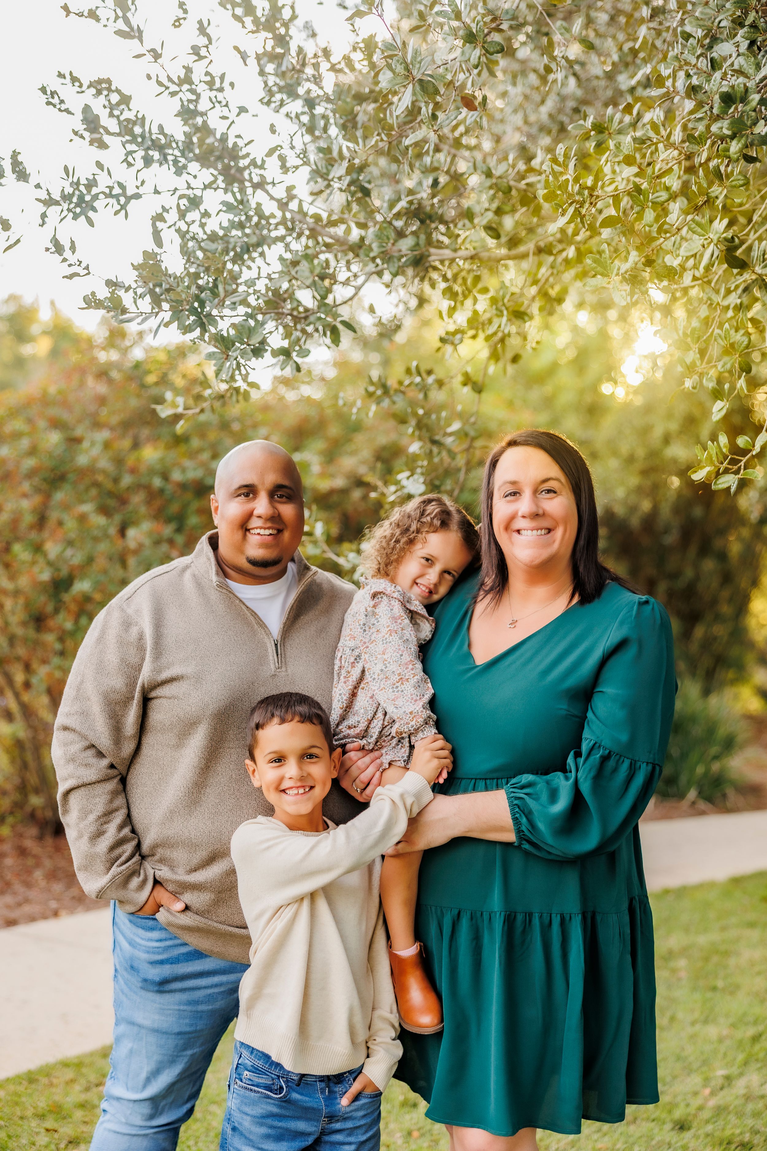 A family of three wearing coordinated casual outfits poses together outdoors during golden hour among olive trees.