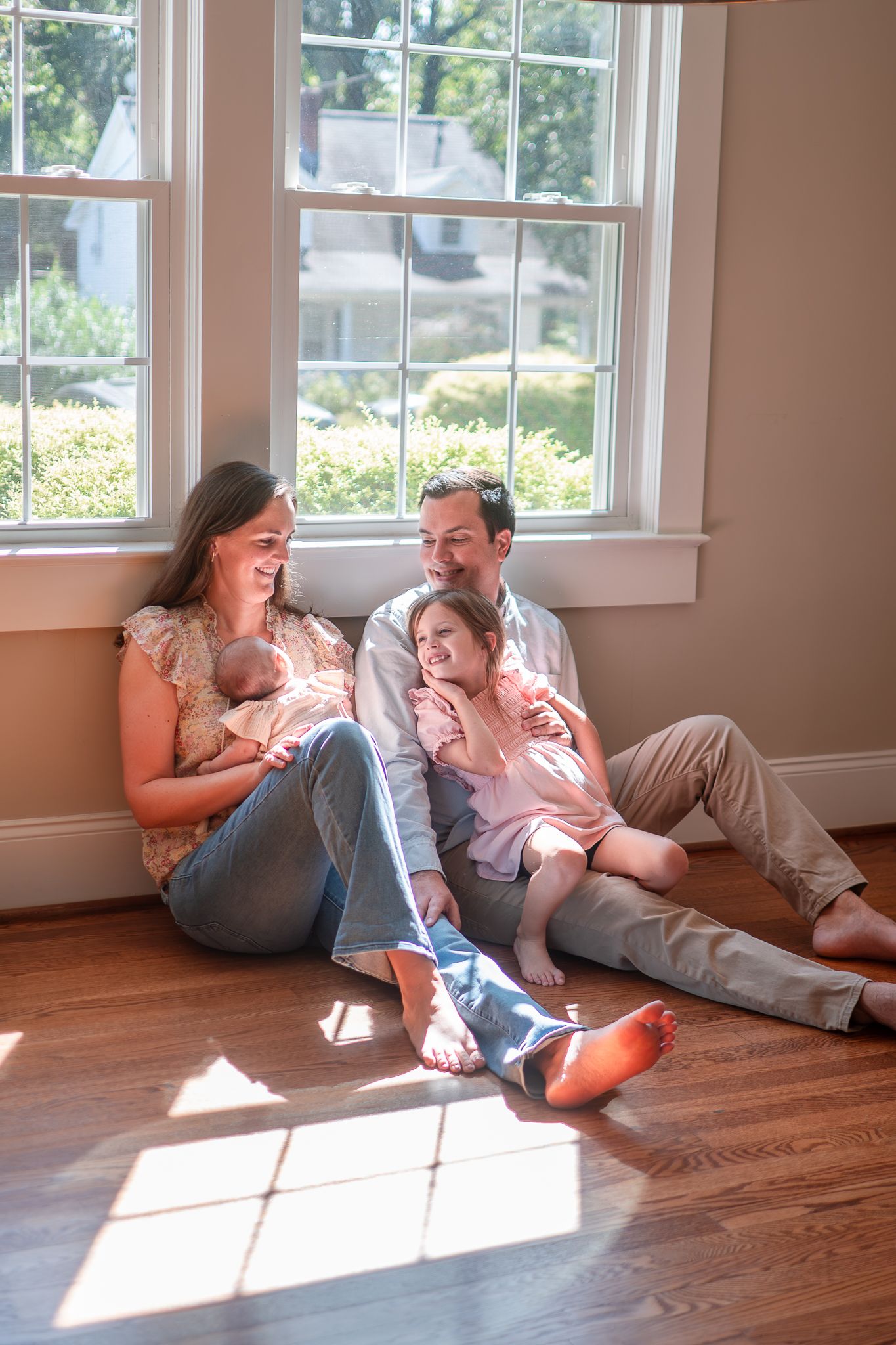 A family shares a playful moment while sitting on a hardwood floor in a sunlit room with white windows.