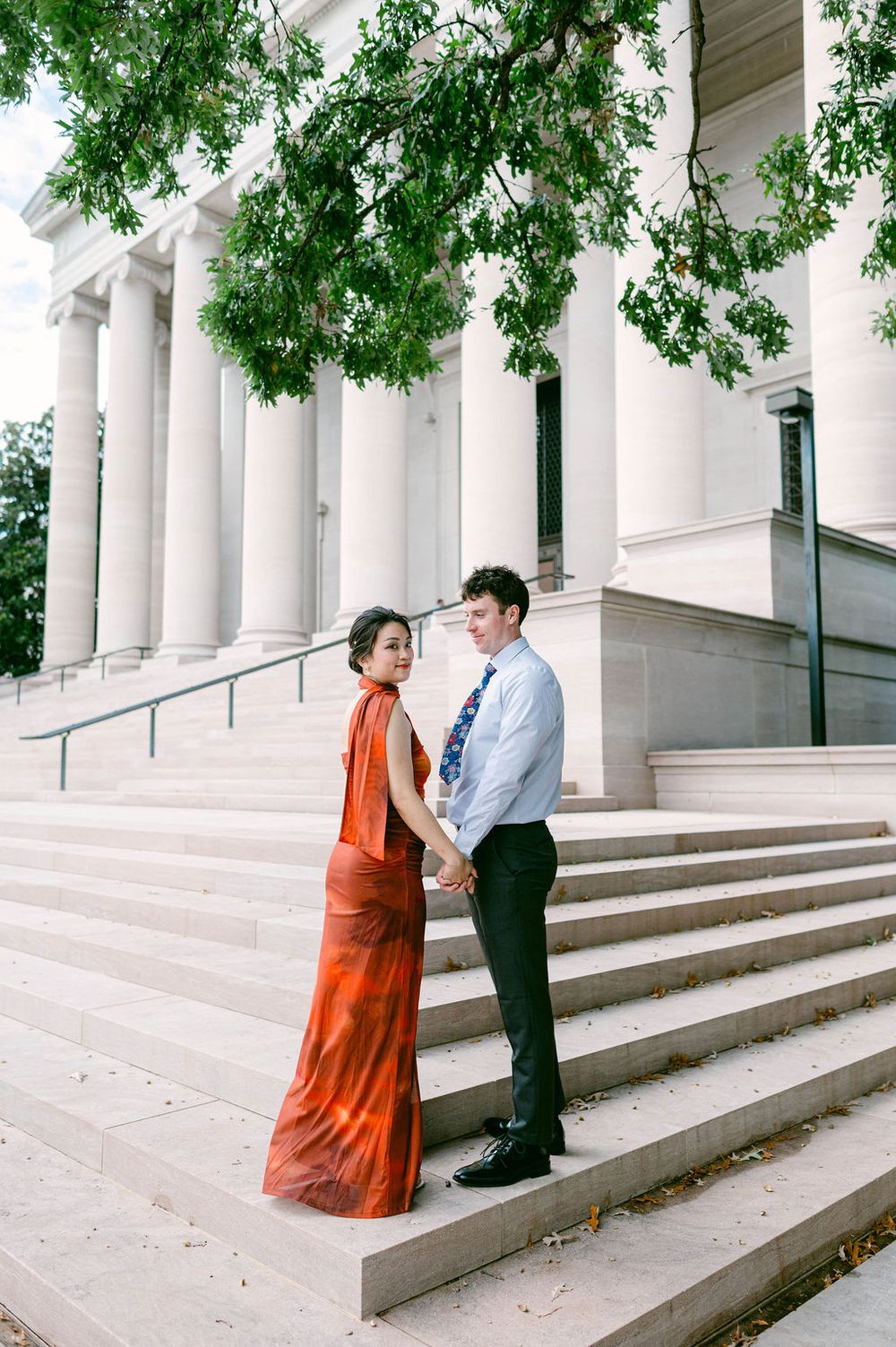 A couple poses on elegant white stone steps with columns, the person in a flowing orange dress creating a striking contrast.