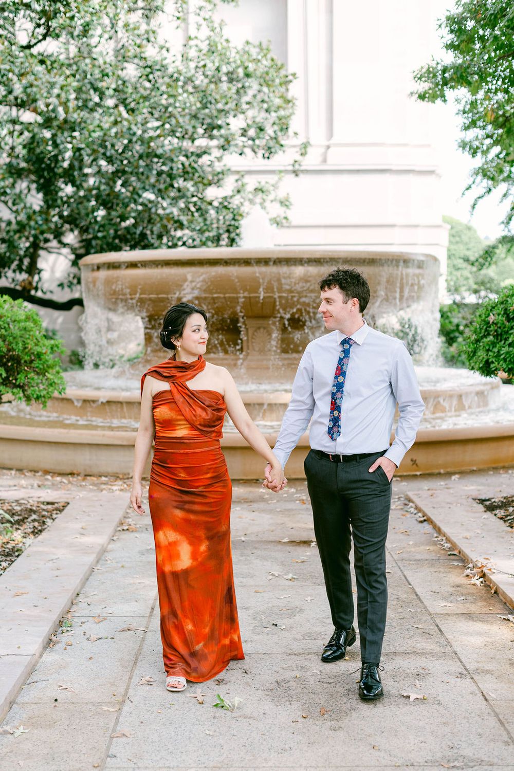 A couple in formal attire dances playfully on a garden path surrounded by greenery.