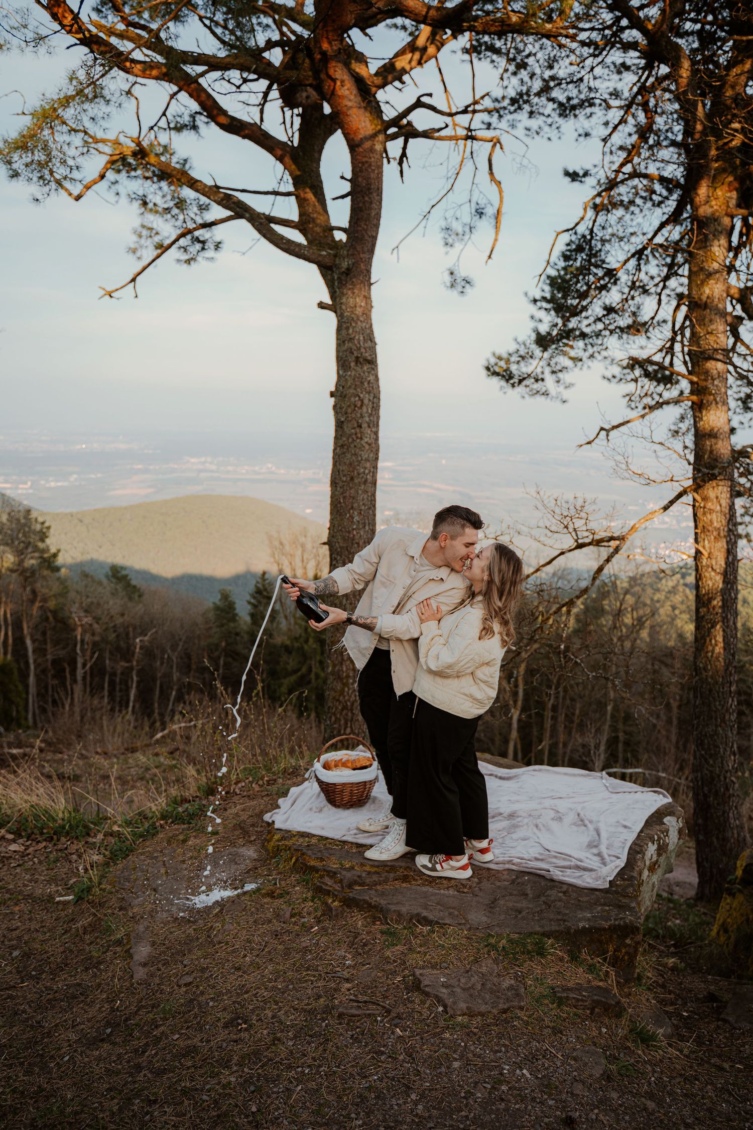 Couple embracing on a hilltop at sunset with tall pine trees framing the romantic mountain landscape.