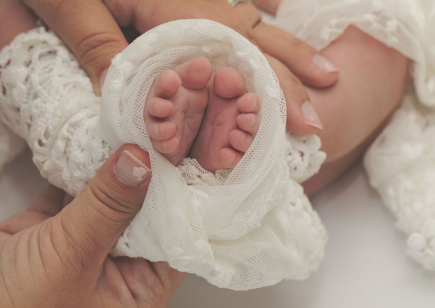 Tiny newborn baby feet wrapped in delicate white lace fabric against a soft pink background.