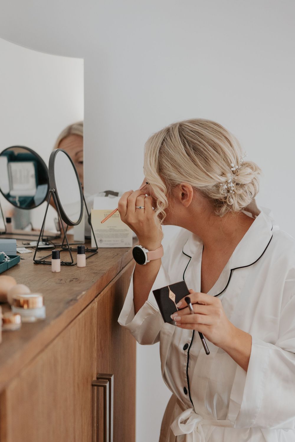 Blonde bride in white pajamas applying makeup at wooden vanity with round mirror.