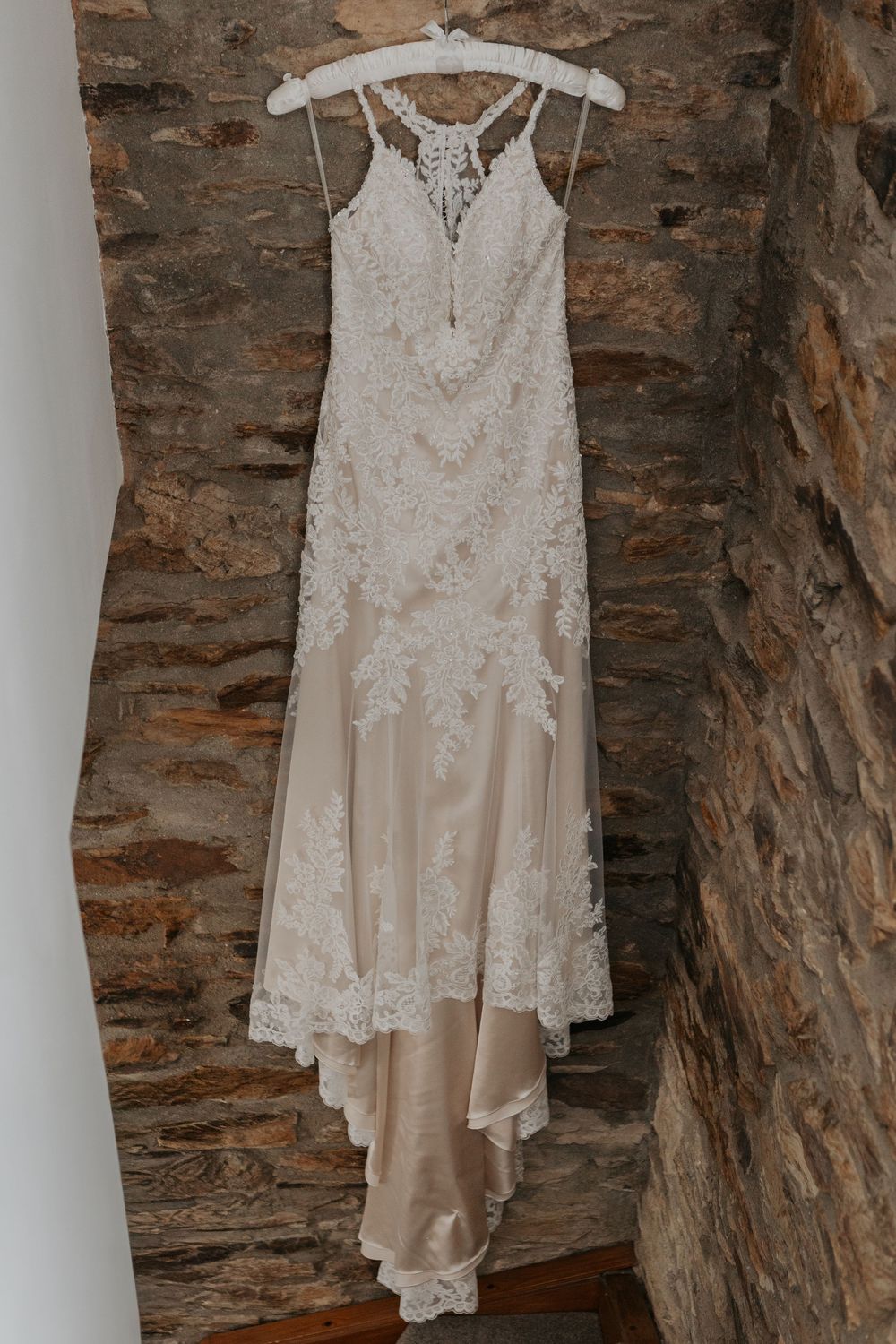Elegant lace wedding dress with champagne underlay hanging on a white hanger against a rustic stone wall.