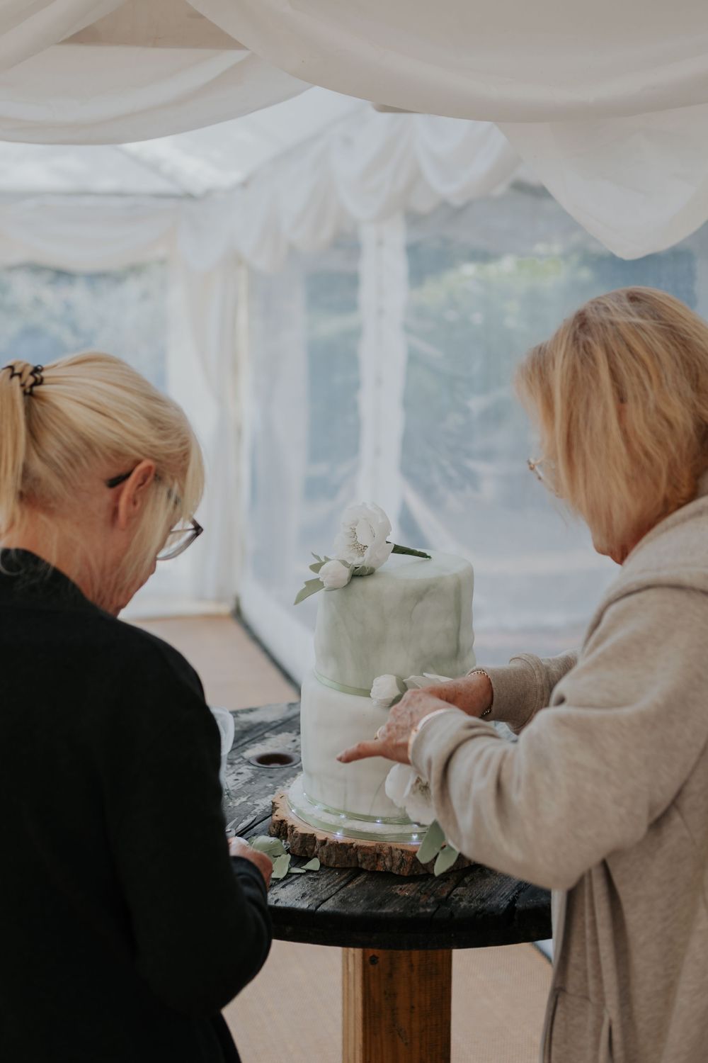 Two women examining a two-tier marble fondant wedding cake decorated with white flowers on a wooden slice stand.