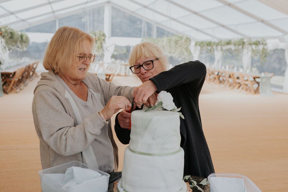 Two women cutting a elegant white tiered wedding cake under a decorated tent.